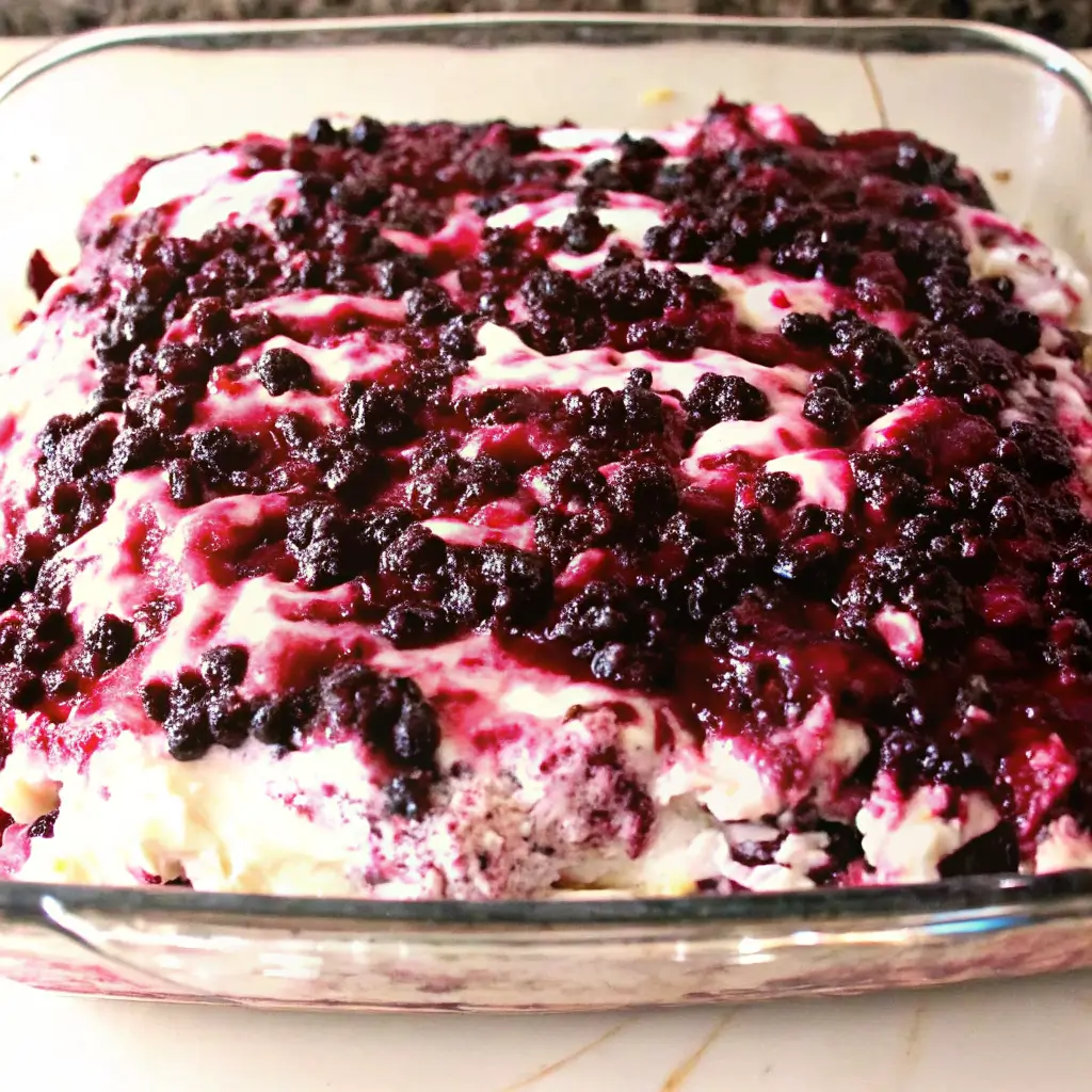 Black Raspberry Cake in a glass baking dish, fresh out of the oven or cooling. The top of the cake is generously layered with black raspberries, which have released their juices during baking, creating deep red-purple swirls across a creamy white layer—likely made of cream cheese or a similar filling. The cake's texture looks soft and rich, with parts of the cake base visible beneath the berry topping. The vibrant contrast between the dark berries and creamy surface gives the cake a rustic, homemade charm perfect for potlucks, family gatherings, or summer desserts.