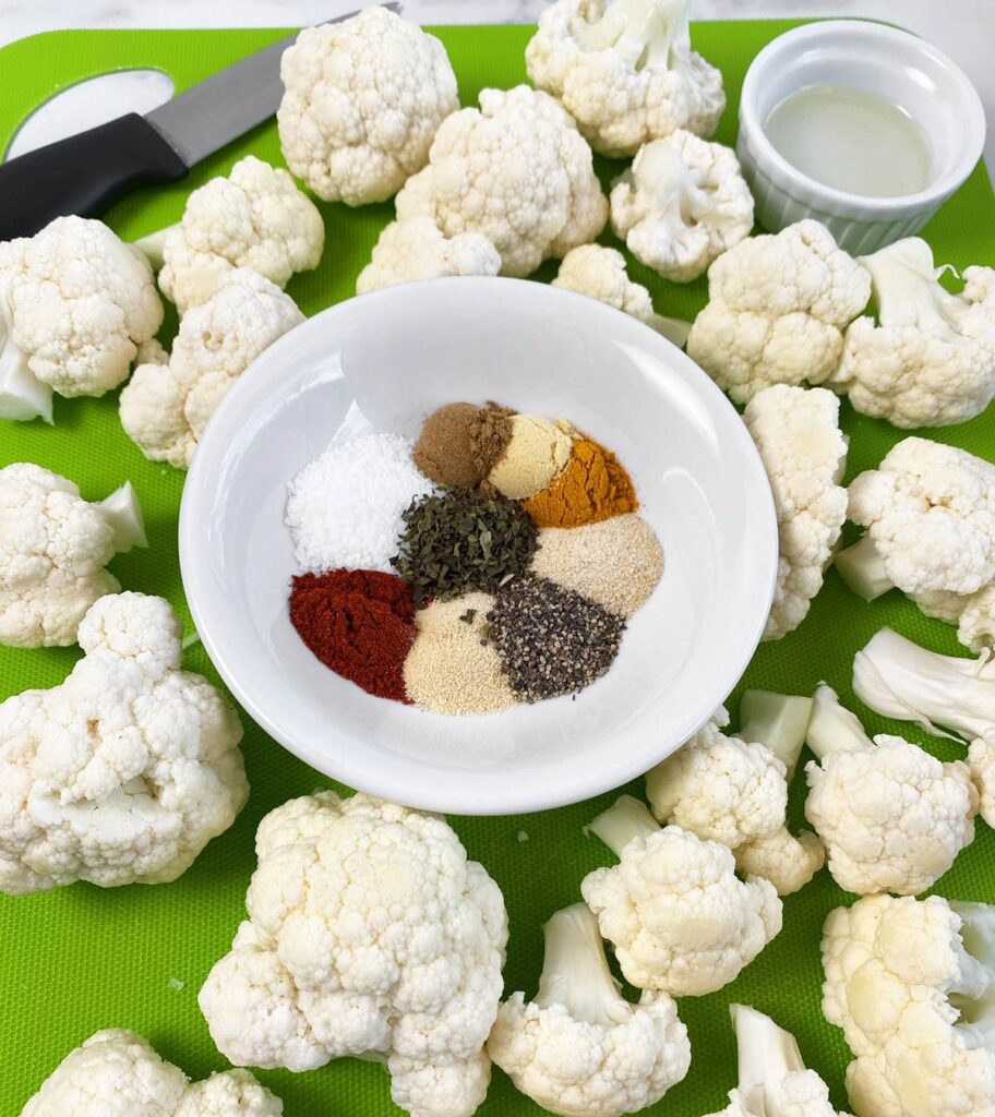Fresh cauliflower florets arranged around a white bowl containing various colorful spices and seasonings on a green cutting mat
